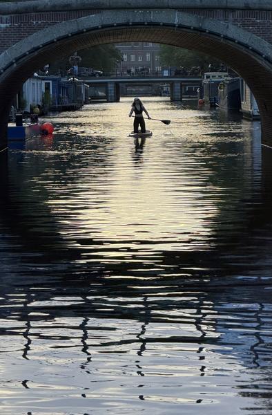 A lone figure is captured stand-up paddleboarding on the tranquil waters of the Amstel canal in Weesperbuurt, Amsterdam, bathed in the warm glow of the evening sun